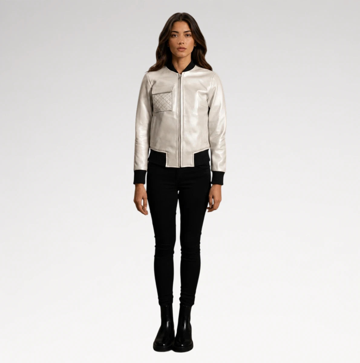 A woman modeling a shiny silver bomber jacket with black trim, paired with black skinny pants and black boots, against a light gray studio background.