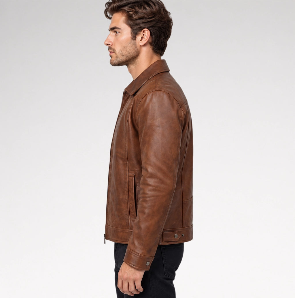 A man wearing a brown leather jacket over a beige t-shirt and dark pants, standing against a light gray studio background.