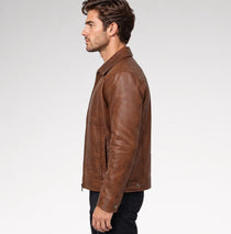 A man wearing a brown leather jacket over a beige t-shirt and dark pants, standing against a light gray studio background.