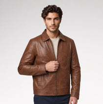 A man wearing a brown leather jacket over a beige t-shirt and dark pants, standing against a light gray studio background.