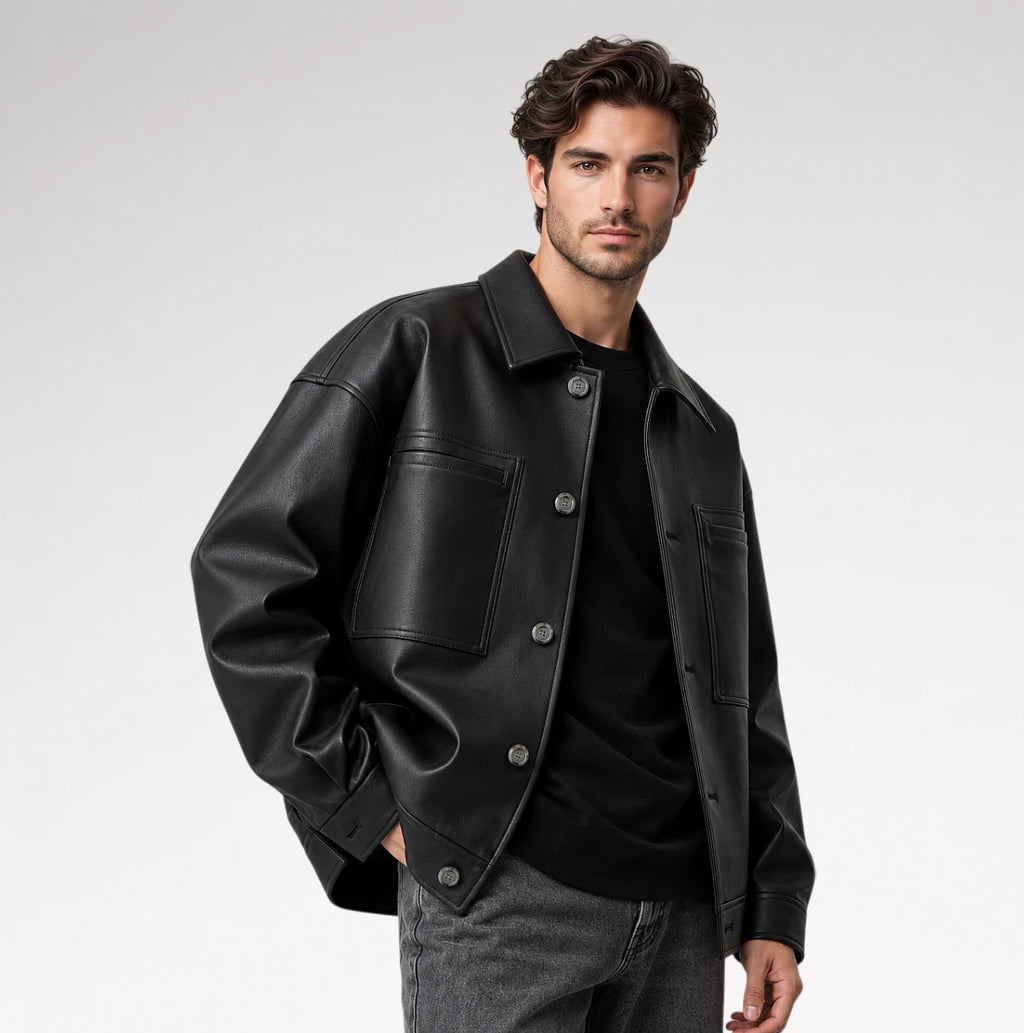 A man wearing a black leather jacket over a black shirt and gray jeans, photographed against a clean light gray studio background.
