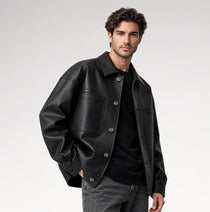 A man wearing a black leather jacket over a black shirt and gray jeans, photographed against a clean light gray studio background.
