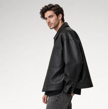 A man wearing a black leather jacket, styled with a black shirt and gray jeans, photographed from a side profile against a light gray studio background.