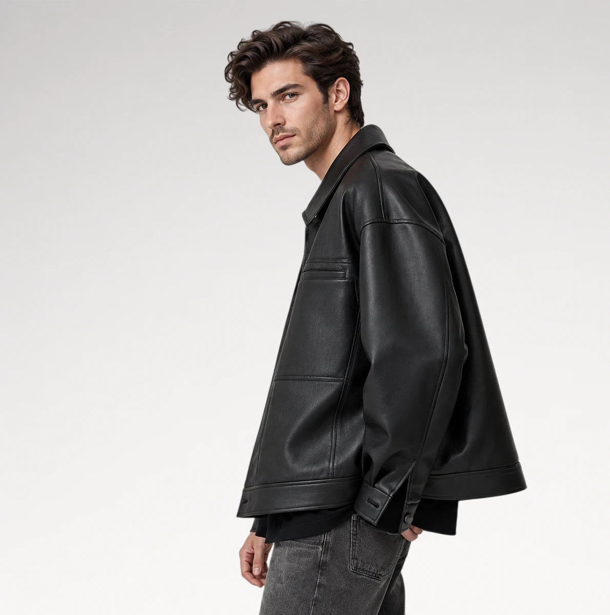 A man wearing a black leather jacket, styled with a black shirt and gray jeans, photographed from a side profile against a light gray studio background.
