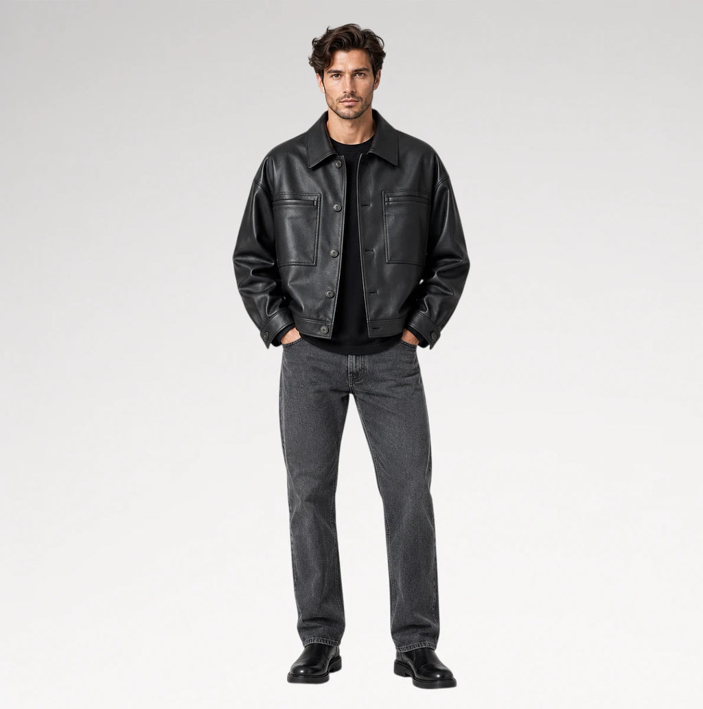 A male model stands against a light gray studio background wearing a black leather jacket over a black shirt, dark gray jeans, and black shoes.