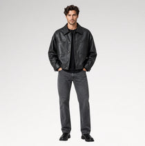 A male model stands against a light gray studio background wearing a black leather jacket over a black shirt, dark gray jeans, and black shoes.