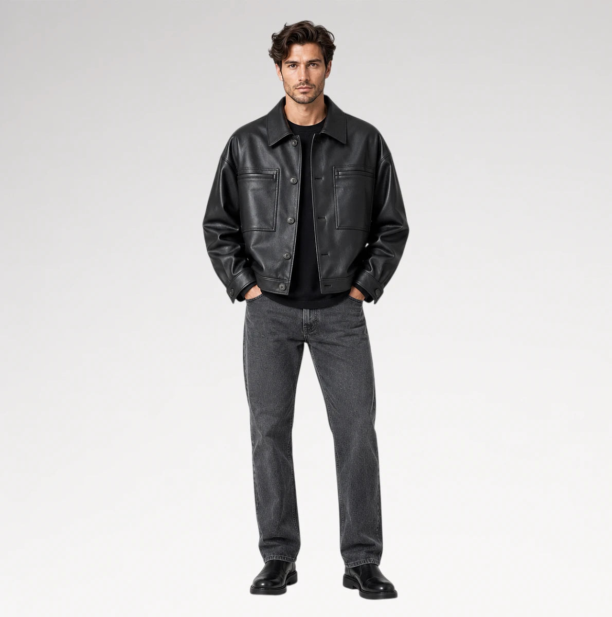 A male model stands against a light gray studio background wearing a black leather jacket over a black shirt, dark gray jeans, and black shoes.