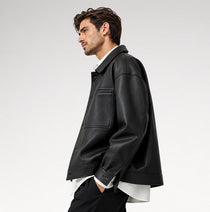 A man in side profile wearing a black leather jacket over a white collared shirt and black trousers, photographed against a clean light gray studio background.
