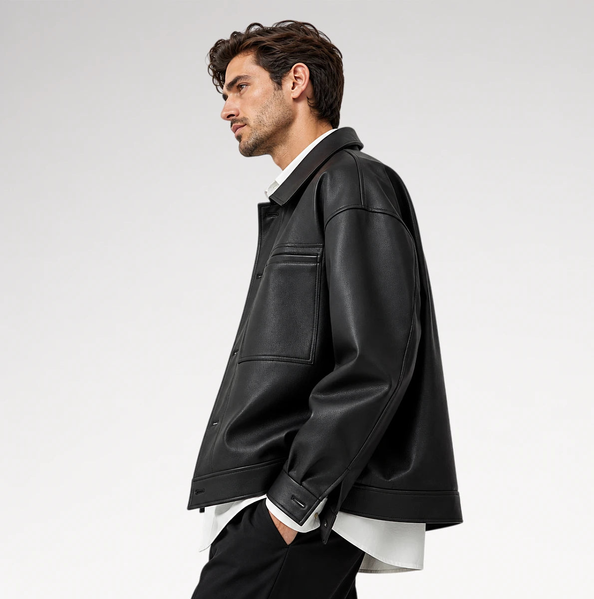 A man in side profile wearing a black leather jacket over a white collared shirt and black trousers, photographed against a clean light gray studio background.