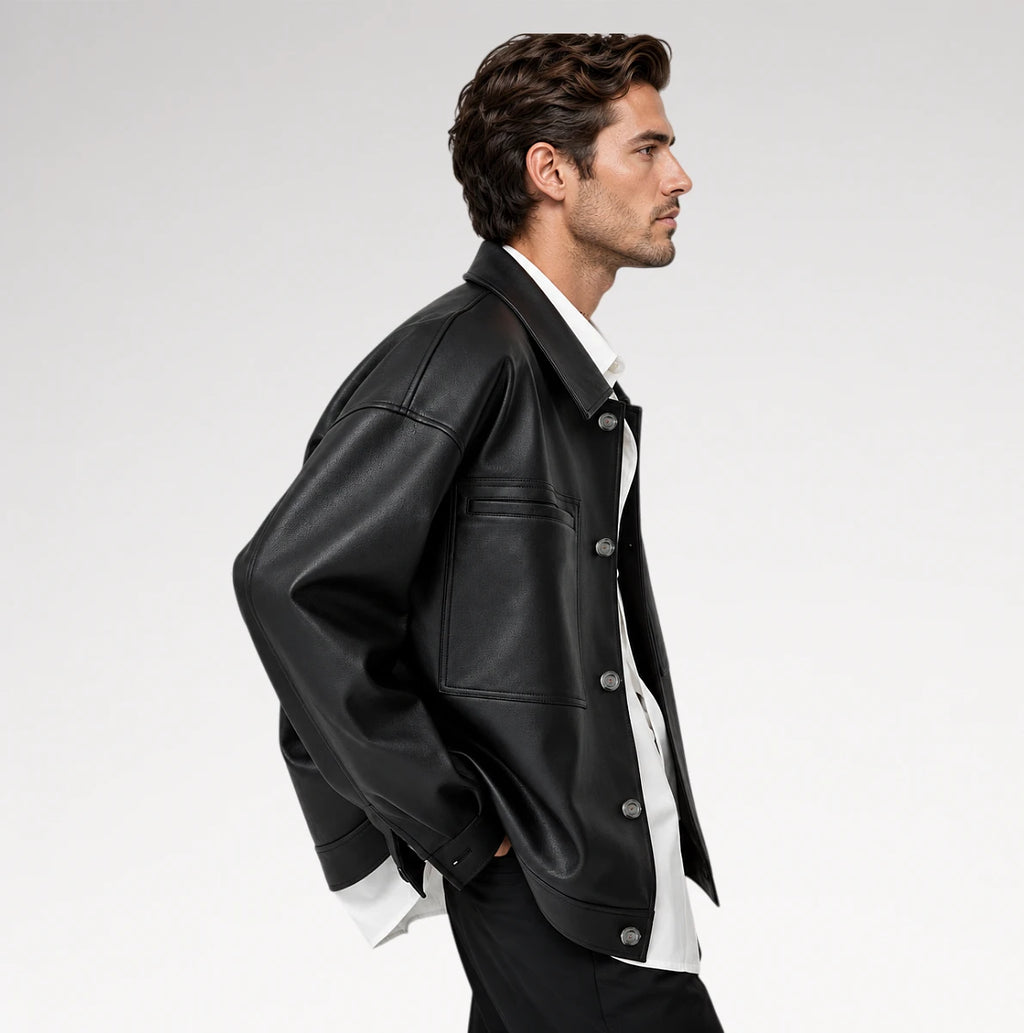 A man in profile wearing a black leather jacket over a white collared shirt and black trousers, photographed against a clean light gray studio background.