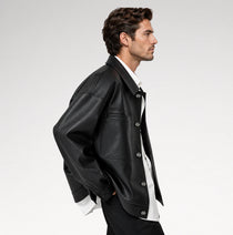 A man in profile wearing a black leather jacket over a white collared shirt and black trousers, photographed against a clean light gray studio background.