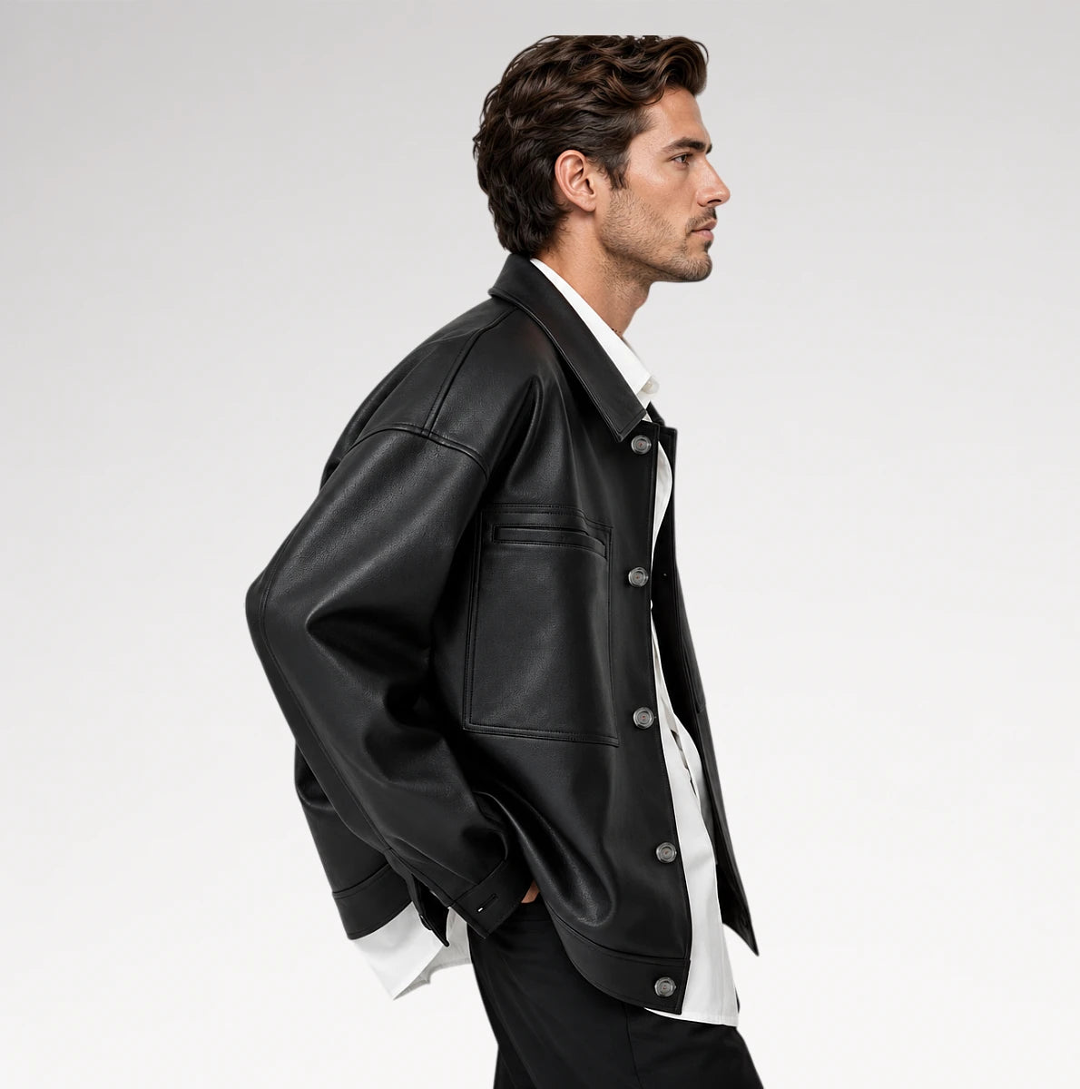 A man in profile wearing a black leather jacket over a white collared shirt and black trousers, photographed against a clean light gray studio background.