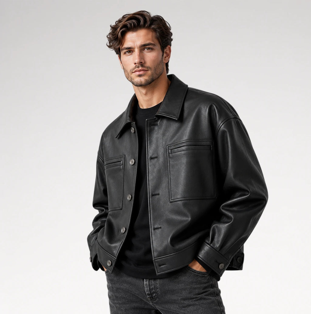 A man wearing a black leather jacket over a black shirt and dark jeans, standing against a light gray studio background.