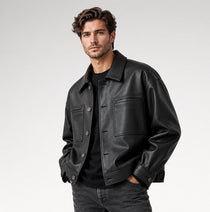 A man wearing a black leather jacket over a black shirt and dark jeans, standing against a light gray studio background.