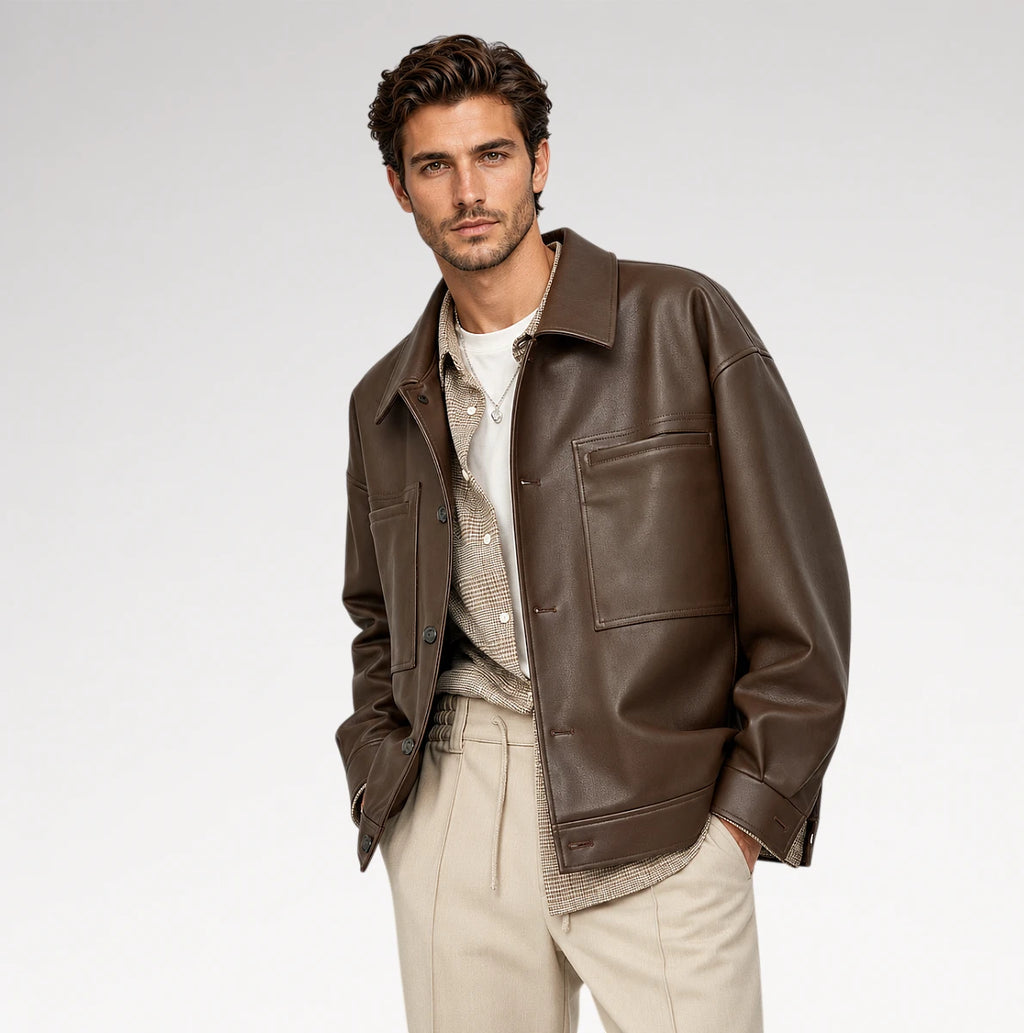 A man wearing a dark brown leather jacket over a beige knit sweater and white t-shirt, paired with beige trousers, photographed against a light gray studio background.