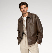 A man wearing a dark brown leather jacket over a beige knit sweater and white t-shirt, paired with beige trousers, photographed against a light gray studio background.
