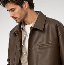 A close-up of a man wearing a brown leather jacket over a beige patterned shirt and white t-shirt, photographed against a light gray studio background.