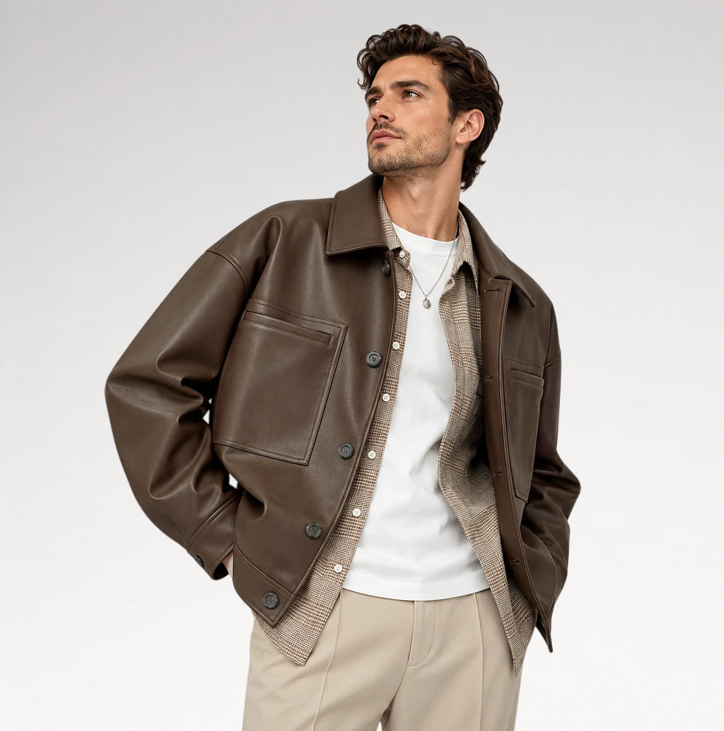 A man wearing a brown leather jacket over a white t-shirt and beige knit sweater, paired with beige trousers, photographed against a light gray studio background.