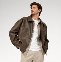 A man wearing a brown leather jacket over a white t-shirt and beige knit sweater, paired with beige trousers, photographed against a light gray studio background.