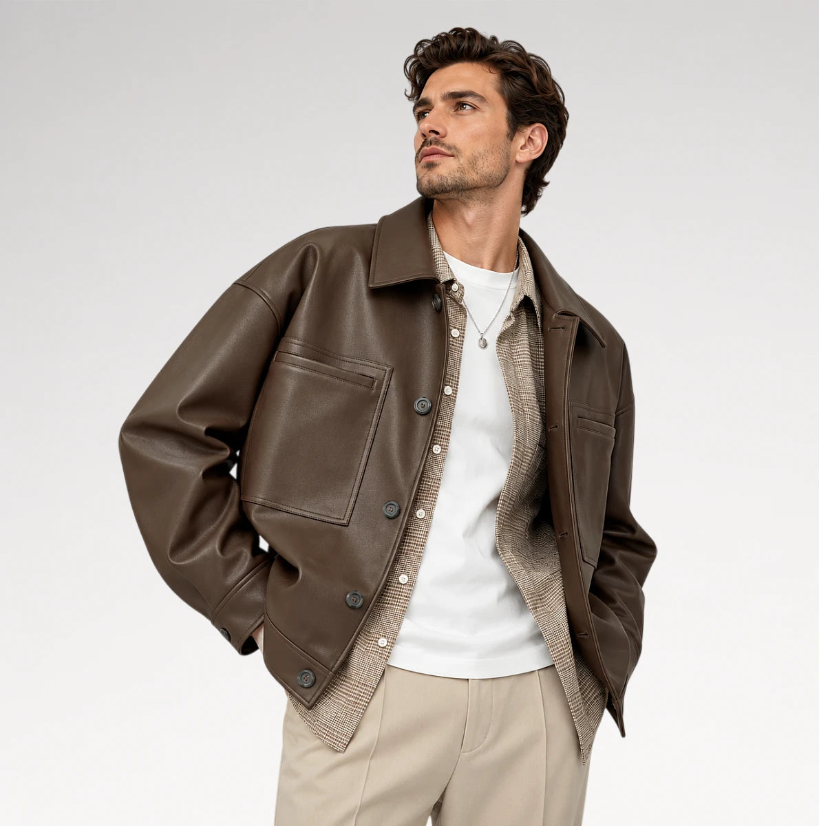 A man wearing a brown leather jacket over a white t-shirt and beige knit sweater, paired with beige trousers, photographed against a light gray studio background.