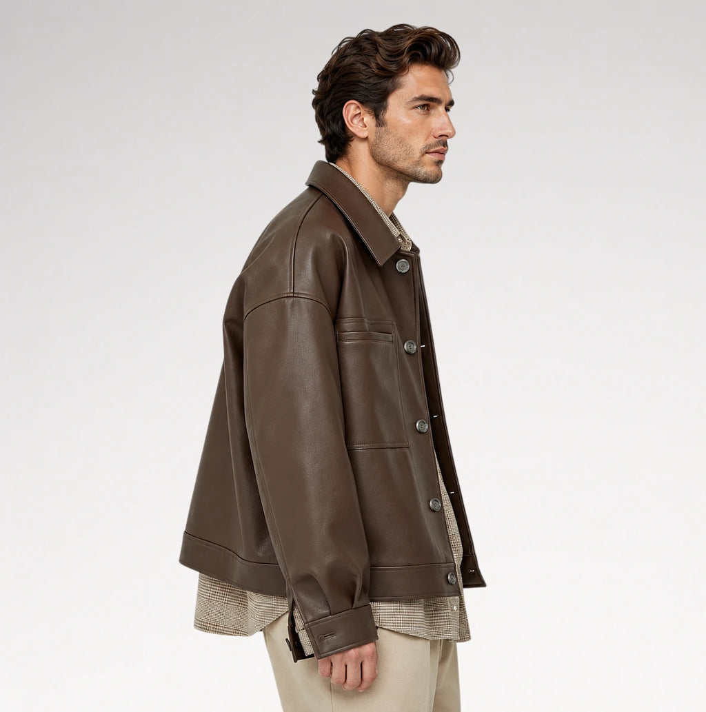 A man in side profile wearing a dark brown leather jacket over a beige shirt and beige trousers, photographed against a clean light gray studio background.
