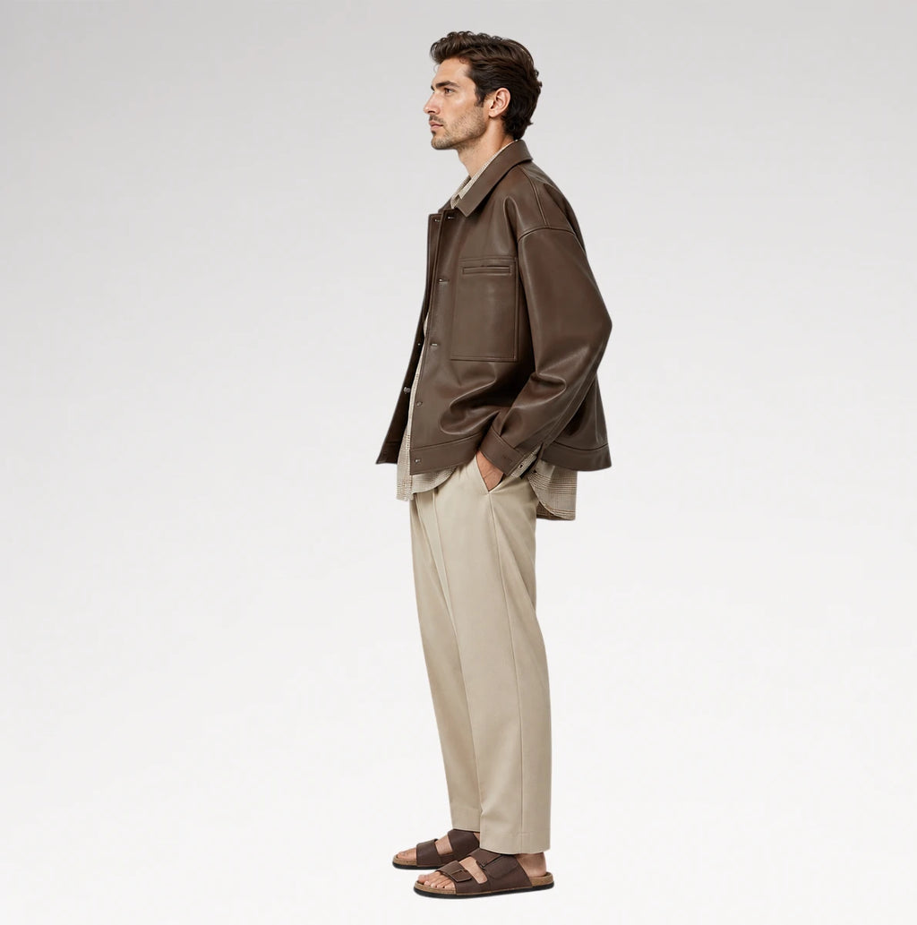 A man in side profile wearing a brown leather jacket, beige trousers, and brown sandals, standing against a clean white studio background.