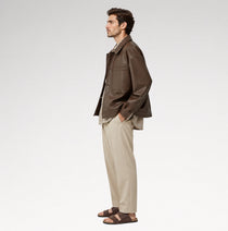 A man in side profile wearing a brown leather jacket, beige trousers, and brown sandals, standing against a clean white studio background.