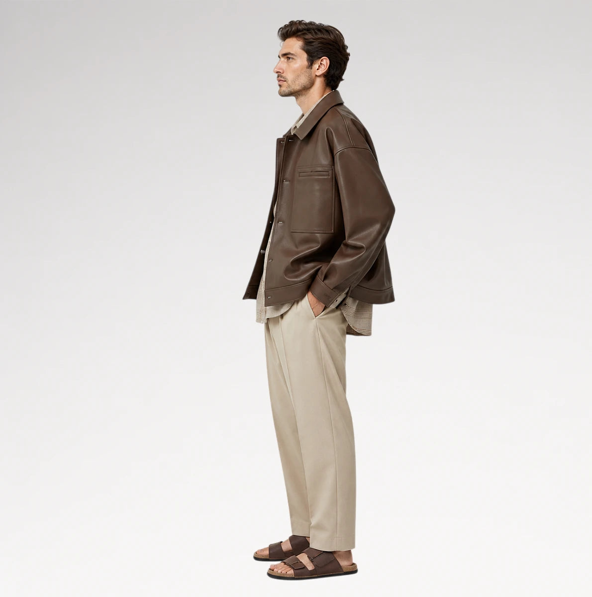 A man in side profile wearing a brown leather jacket, beige trousers, and brown sandals, standing against a clean white studio background.