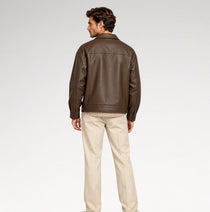 A man shown from the back wearing a dark brown leather jacket and beige trousers, standing against a light gray studio background.