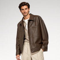 A male model wearing a dark brown leather jacket over a beige knit sweater and white t-shirt, paired with beige trousers, photographed against a light gray studio background.