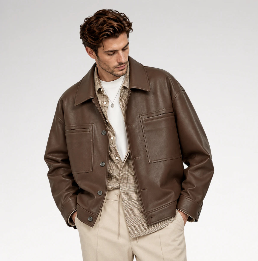 A man wearing a brown leather jacket over a beige patterned shirt and white t-shirt, paired with beige trousers, photographed against a light gray studio background.