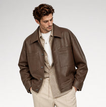 A man wearing a brown leather jacket over a beige patterned shirt and white t-shirt, paired with beige trousers, photographed against a light gray studio background.