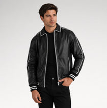 A man wearing a black leather bomber jacket with white contrast piping and striped ribbed cuffs, paired with a black t-shirt and black pants, photographed against a light gray studio background.