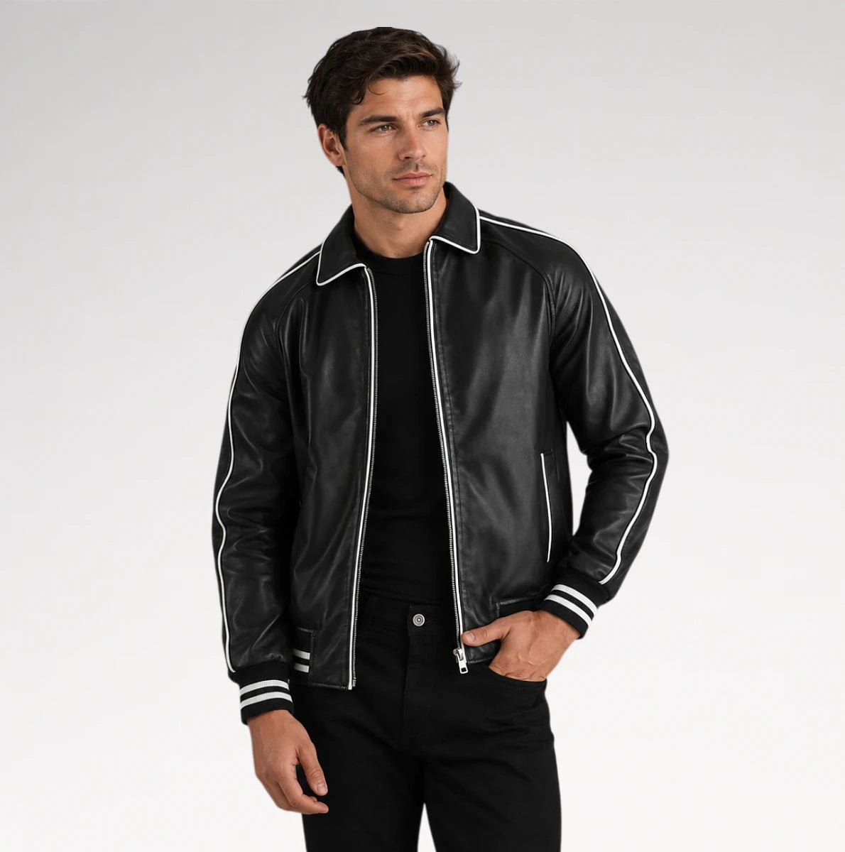 A man wearing a black leather bomber jacket with white contrast piping and striped ribbed cuffs, paired with a black t-shirt and black pants, photographed against a light gray studio background.