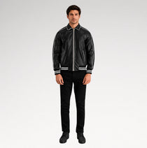 A man modeling a black leather bomber jacket with white contrast stitching and striped ribbed collar, cuffs, and hem, paired with black pants and black shoes, against a light gray studio background.