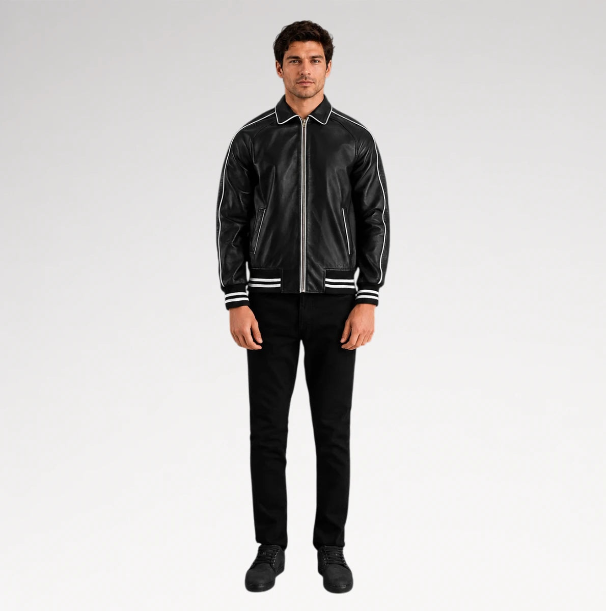 A man modeling a black leather bomber jacket with white contrast stitching and striped ribbed collar, cuffs, and hem, paired with black pants and black shoes, against a light gray studio background.
