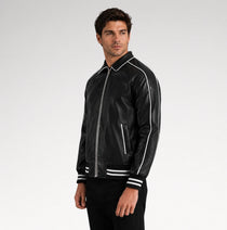 A man wearing a black leather bomber jacket with white contrast stitching and striped ribbed cuffs, standing against a light gray studio background.