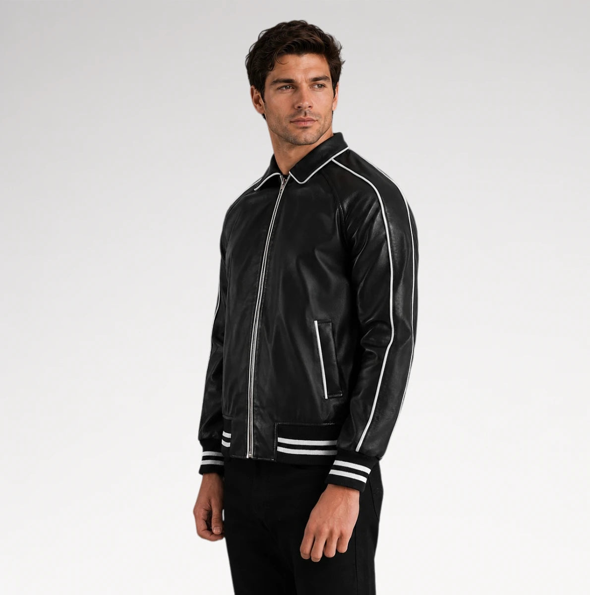 A man wearing a black leather bomber jacket with white contrast stitching and striped ribbed cuffs, standing against a light gray studio background.