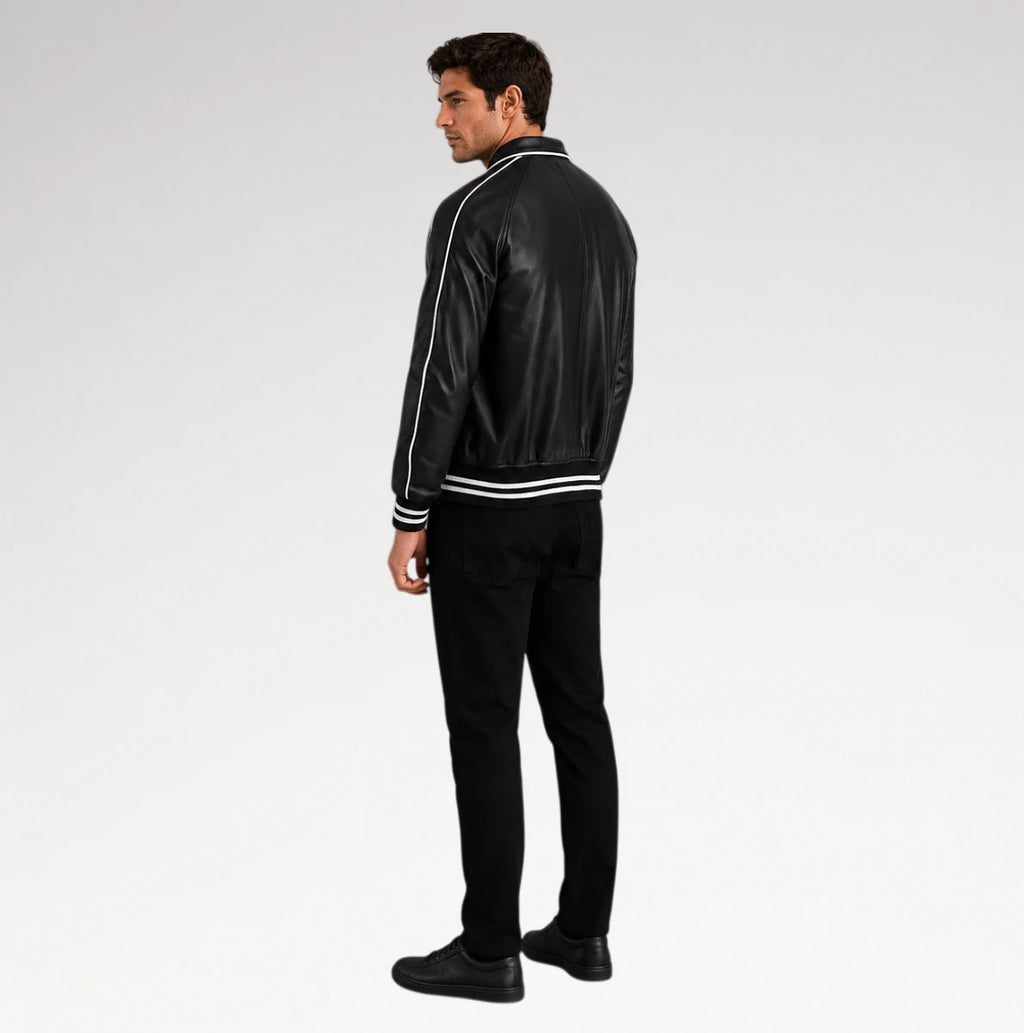 A man wearing a black faux leather bomber jacket with white contrast stitching and ribbed trim, paired with black pants and shoes, shown from a side-back angle against a light gray studio background.