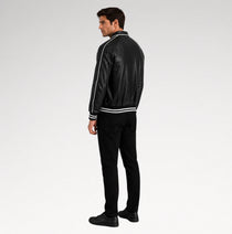 A man wearing a black faux leather bomber jacket with white contrast stitching and ribbed trim, paired with black pants and shoes, shown from a side-back angle against a light gray studio background.