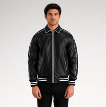 A man wearing a black leather bomber jacket with white contrast piping, a white collar, and white stripes on the cuffs and hem, paired with black pants against a light gray studio background.
