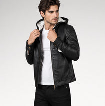 A man wearing a black faux-leather hooded jacket with a white hood lining, styled over a white t-shirt and black jeans, photographed against a light gray studio background.