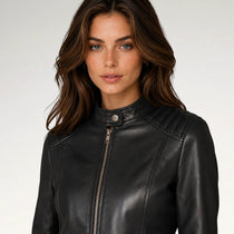 A woman with long brown hair wearing a black leather jacket with a high collar and front zipper, photographed against a light gray studio background.