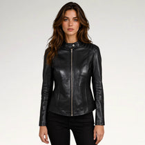 A woman modeling a black faux-leather moto jacket with a silver front zipper and snap collar, paired with black pants, against a plain light gray studio background.
