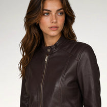 A woman wearing a dark brown faux-leather moto jacket with a snap collar and front zipper, photographed against a light gray studio background.
