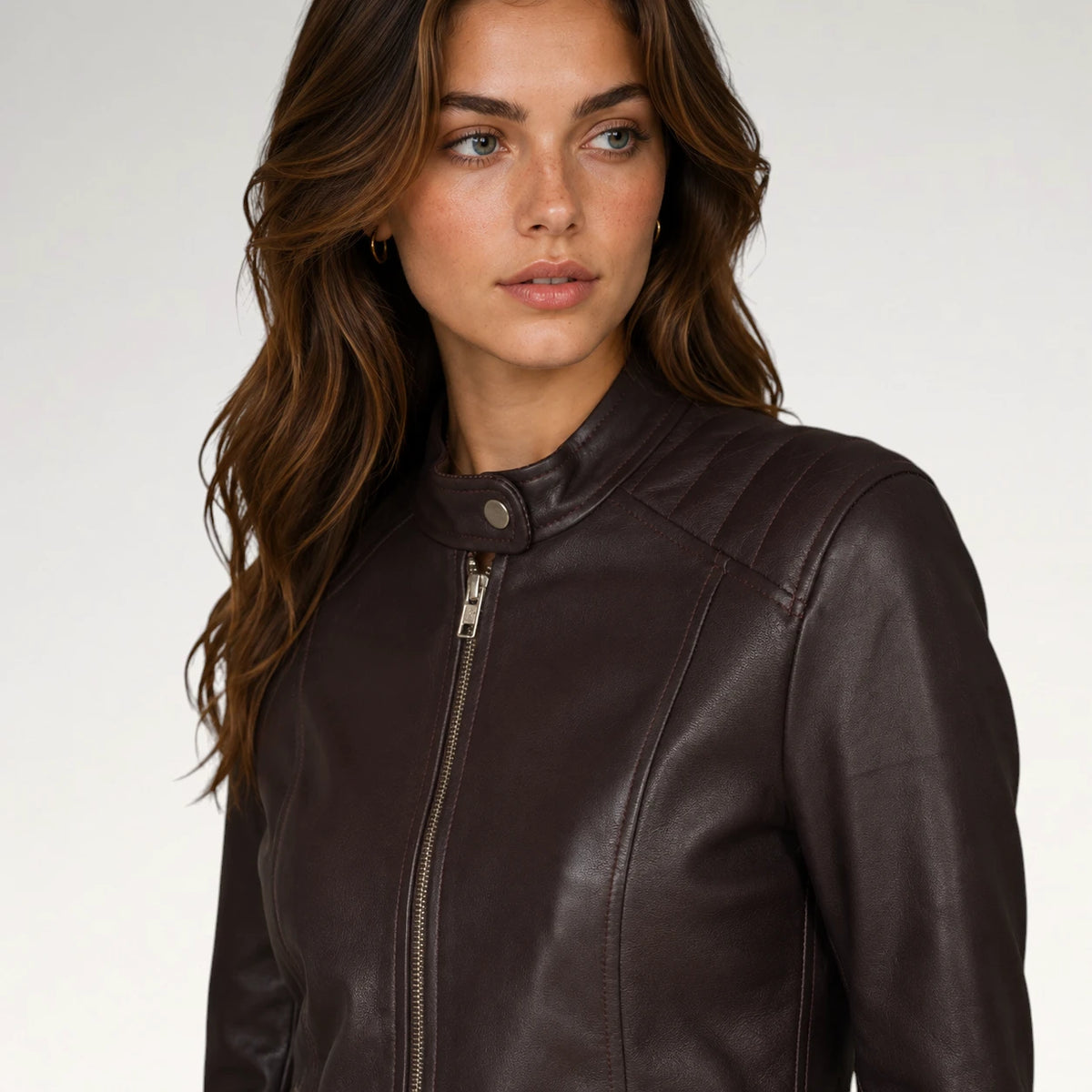 A woman wearing a dark brown faux-leather moto jacket with a snap collar and front zipper, photographed against a light gray studio background.