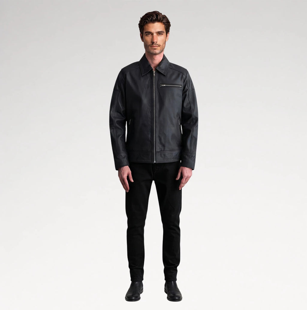 A man modeling a black leather jacket with a zippered chest pocket, paired with black pants and black shoes, against a light gray studio background.