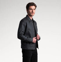 A man wearing a dark gray leather jacket over a black shirt and black pants, standing against a light gray studio background.