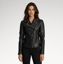 A woman modeling a black leather moto jacket with silver studs and zippers, paired with black pants, against a light gray studio background.
