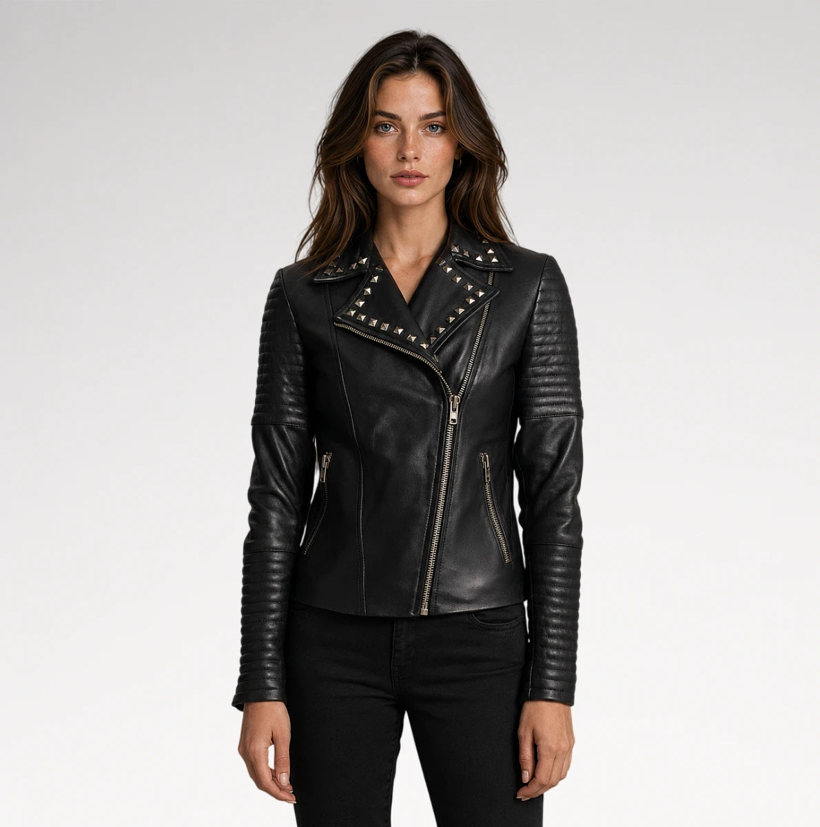 A woman modeling a black leather moto jacket with silver studs and zippers, paired with black pants, against a light gray studio background.